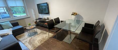Una sala de estar moderna con una mesa de comedor de vidrio, sofás negros, un televisor montado en la pared y vistas al exterior a través de amplios ventanales.