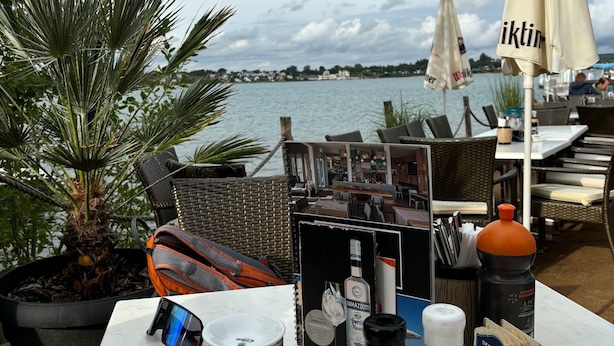 A table with a slice of cheesecake, a glass of beer, a mug of tea, and a bottle of water, with a view of the water and sky in the background. - Holiday Apartment 'Terrassenwohnung' with Private Terrace and Wi-Fi (Rötha)