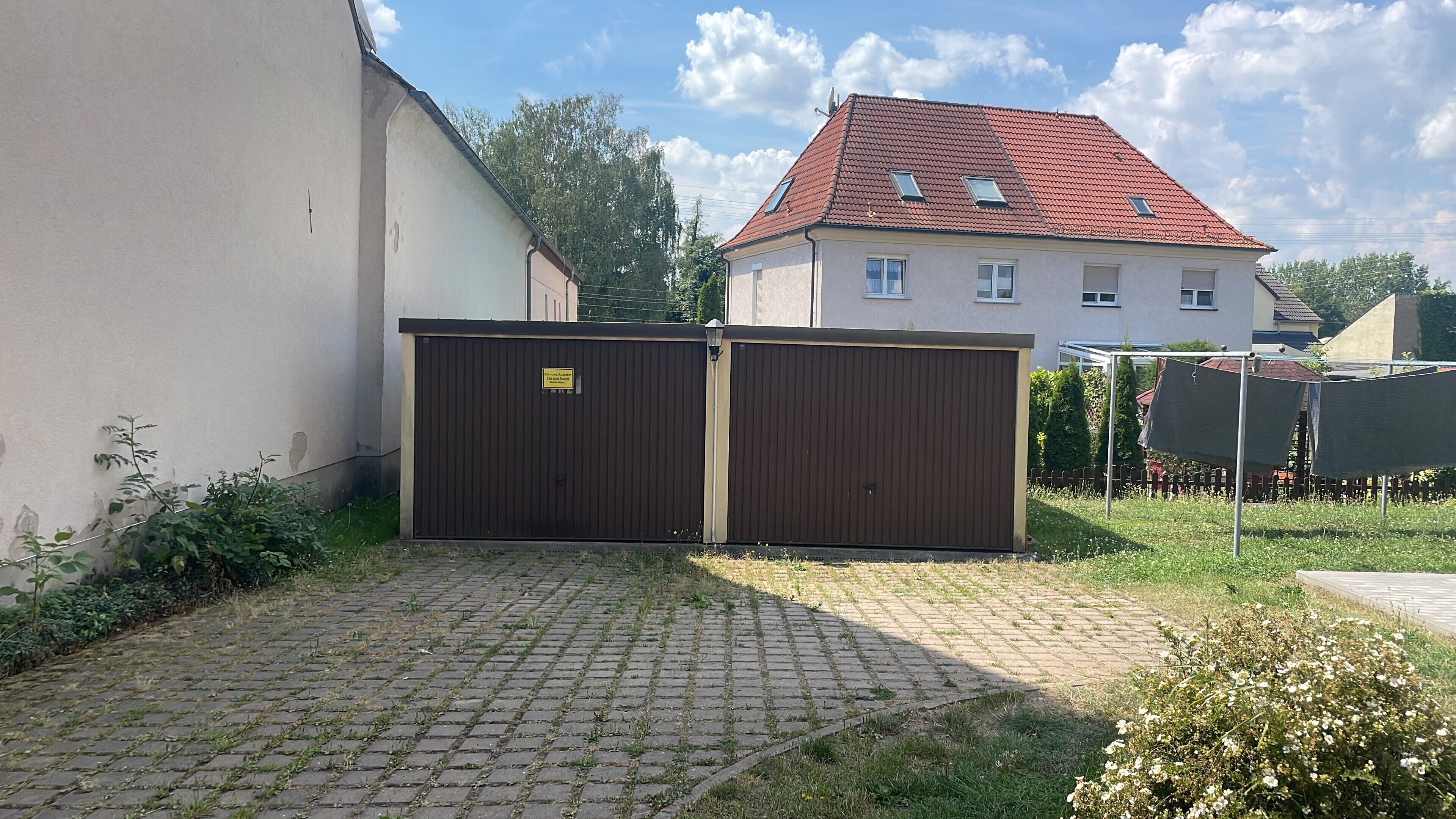 Ein verschlossenes Tor zu einem Wohngebiet, eine gepflasterte Auffahrt führt zu einem Haus mit roten Ziegeldach.