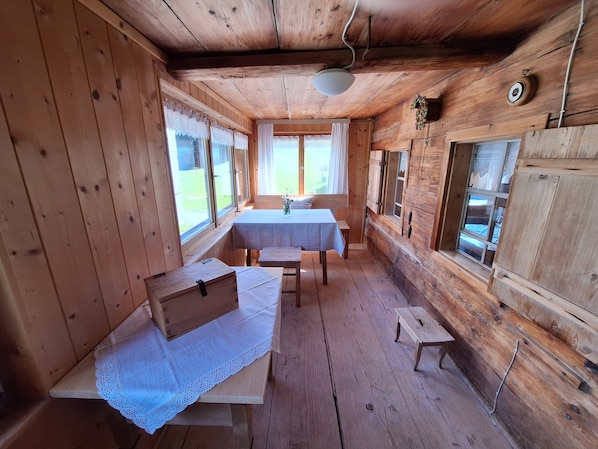 A wooden cabin interior with a dining table, benches, and a window with curtains.