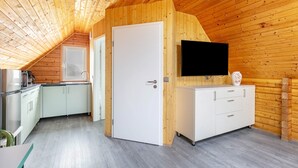 Huone, jossa on kalteva katto, puuseinät, valkoinen ovi, keittiöalue kaapistoineen ja työtasoineen sekä seinälle asennettu litteänäyttöinen televisio.