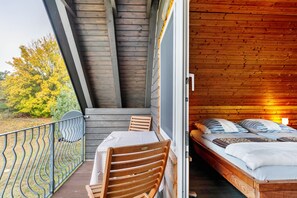 A wooden balcony with a table and chairs, a bed with pillows, and a view of trees.