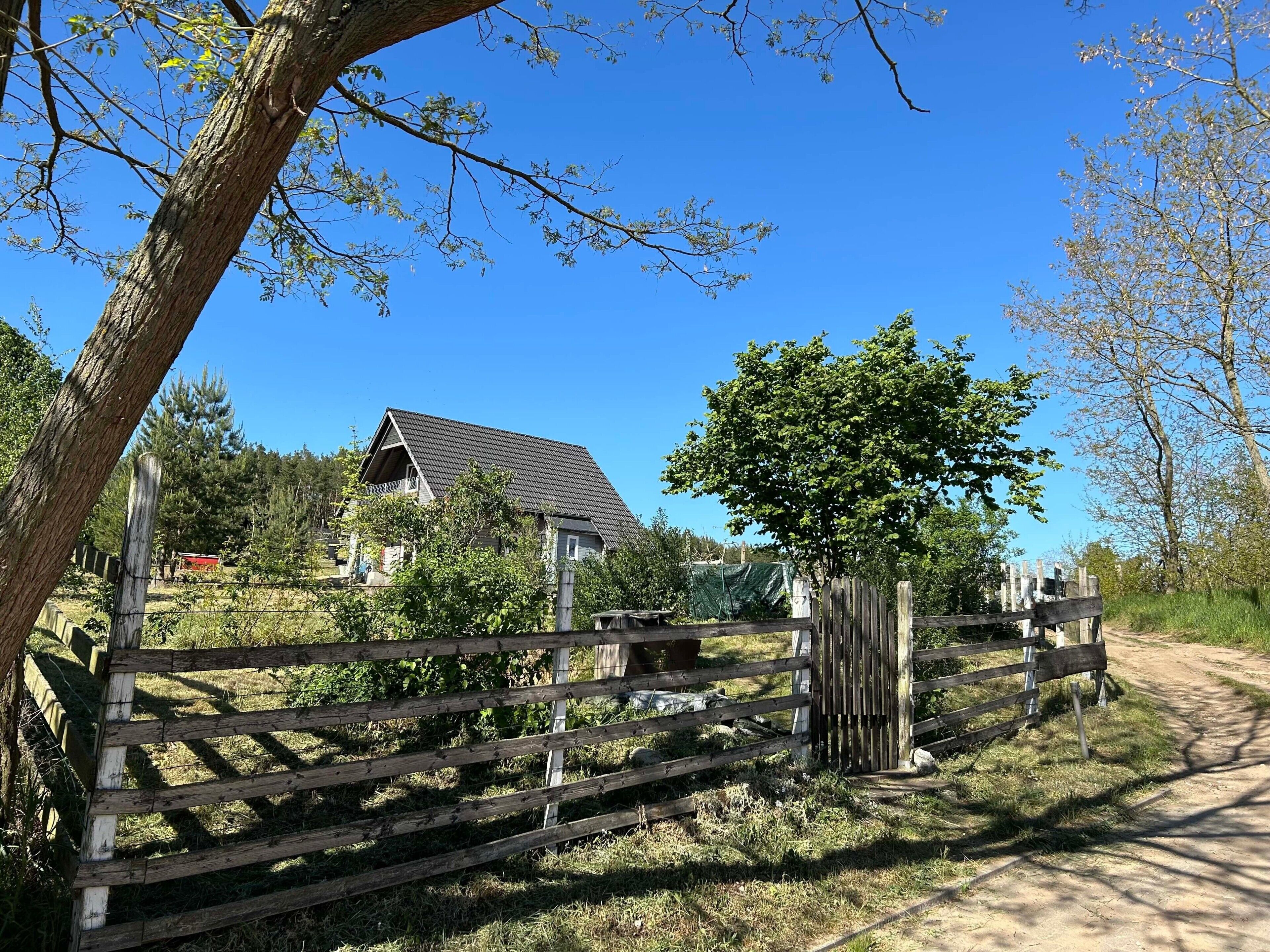 Une maison de campagne avec un toit en pente, entourée d’une clôture en bois et d’arbres.