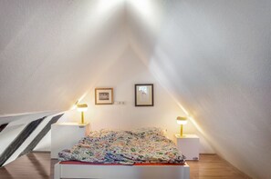 Ein Schlafzimmer mit schrägen Wänden, einem Bett mit floraler Bettdecke, zwei Nachttischlampen und zwei gerahmten Bildern an der Wand.