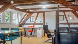 A room with a sloped ceiling, wooden beams, a desk with a chair, a couch, and a window with curtains. - Apartment 'Alte Schmiede Ellrichshausen' with Wi-Fi (Satteldorf)