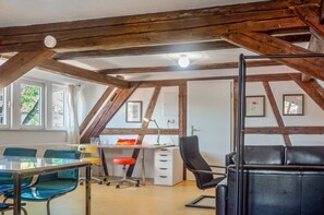 Ein Zimmer mit schrägen Wänden, Holzbalken, einem Schreibtisch mit Stuhl, einer Couch und einem Fenster mit Vorhängen.