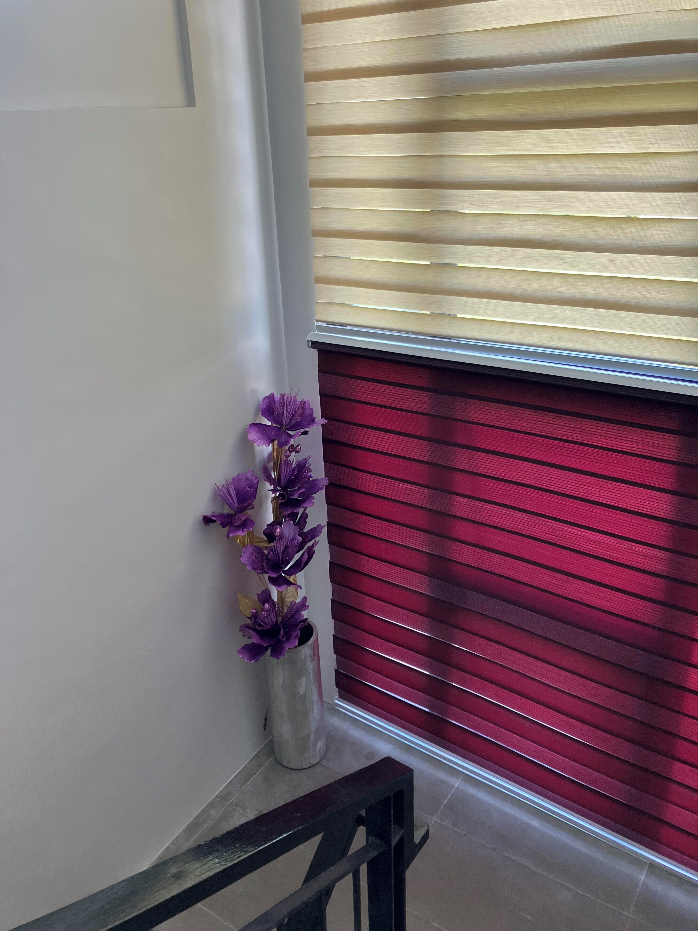 Una escalera con barandilla metálica, pared blanca y una ventana con persianas horizontales.