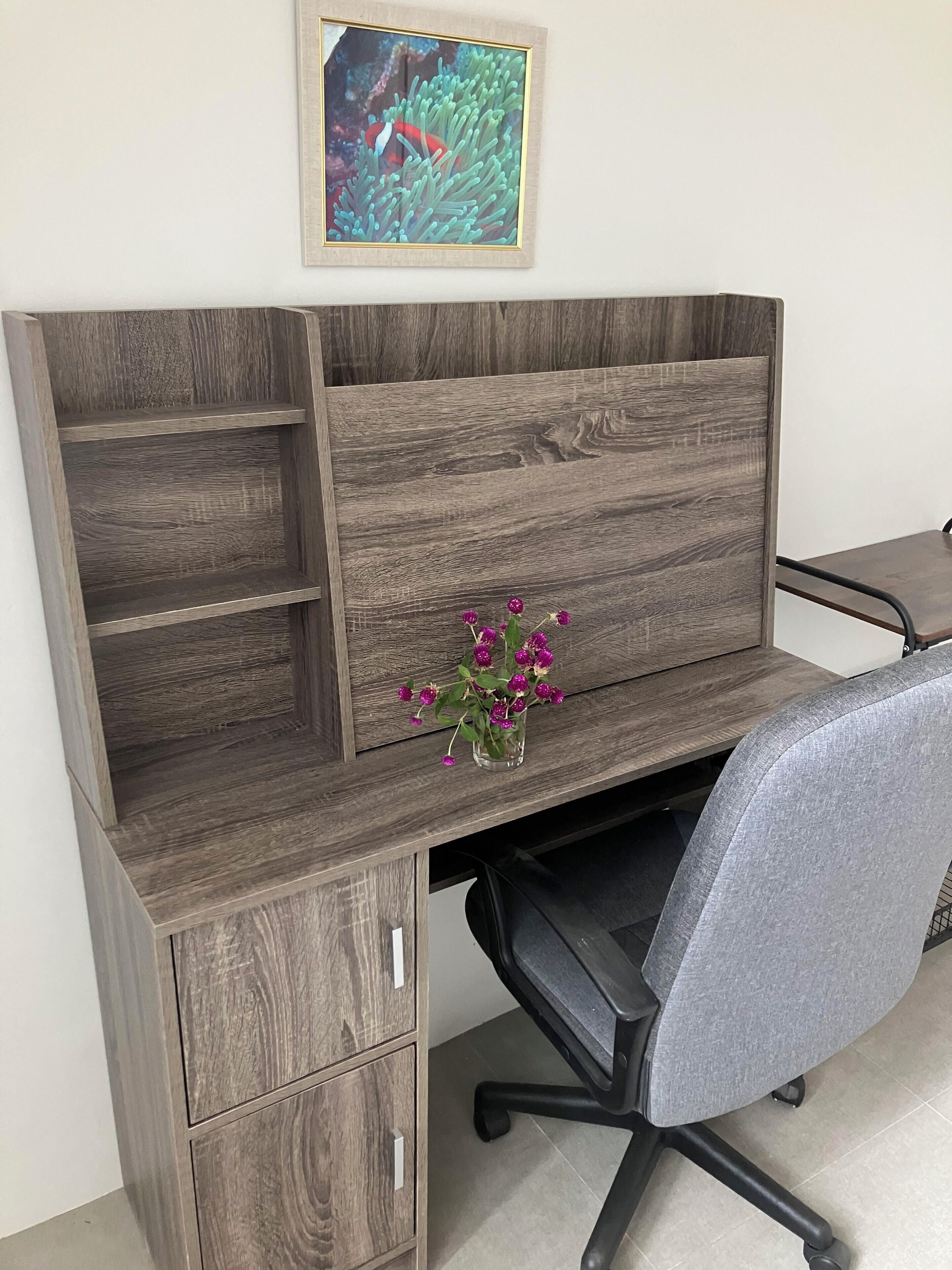A wooden desk with a vase of purple flowers, a chair, and a framed picture on the wall.