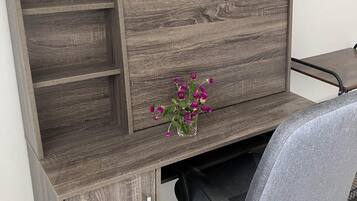 A wooden desk with a vase of purple flowers, a chair, and a framed picture on the wall.