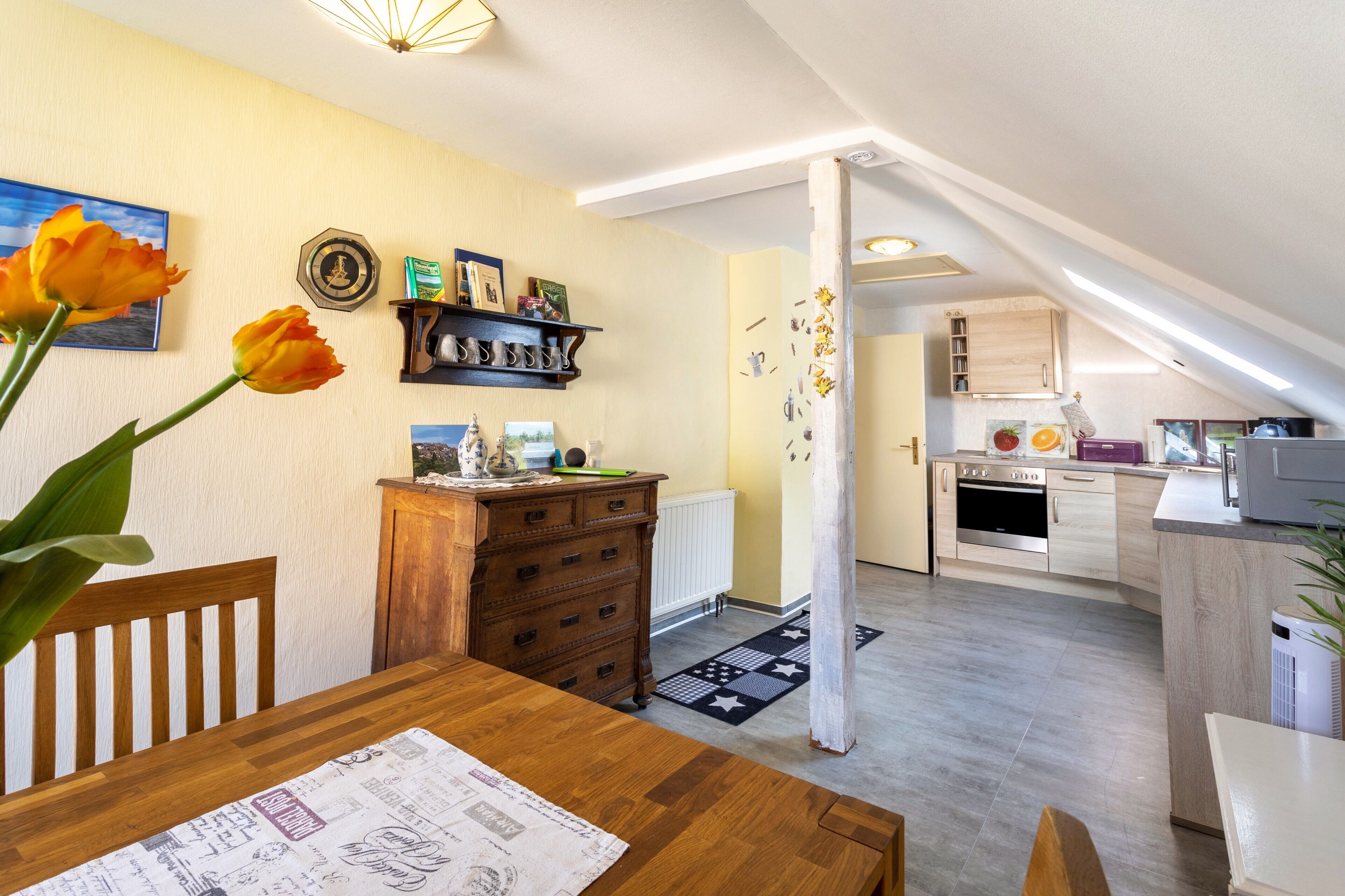 A dining area with a wooden table, chairs, and a vase of flowers. There is a kitchen in the background with modern appliances and wooden cabinets.