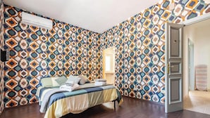 Een slaapkamer met behang met een patroon, een bed met een gestreept dekbed en een open deur naar een andere kamer.