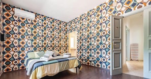 Une chambre avec un papier peint à motifs, un lit avec des oreillers, une table de chevet et une porte ouverte donnant sur une autre pièce.