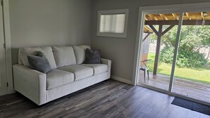 Un salon aux couleurs neutres, avec un canapé gris, une petite fenêtre et une porte-fenêtre coulissante donnant sur une terrasse extérieure.
