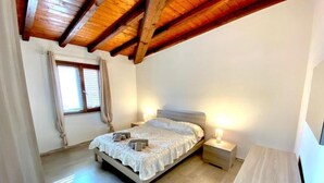 Een slaapkamer met een houten plafond, een bed met een witte dekbedovertrek, twee nachtlampjes en een raam met gordijnen.