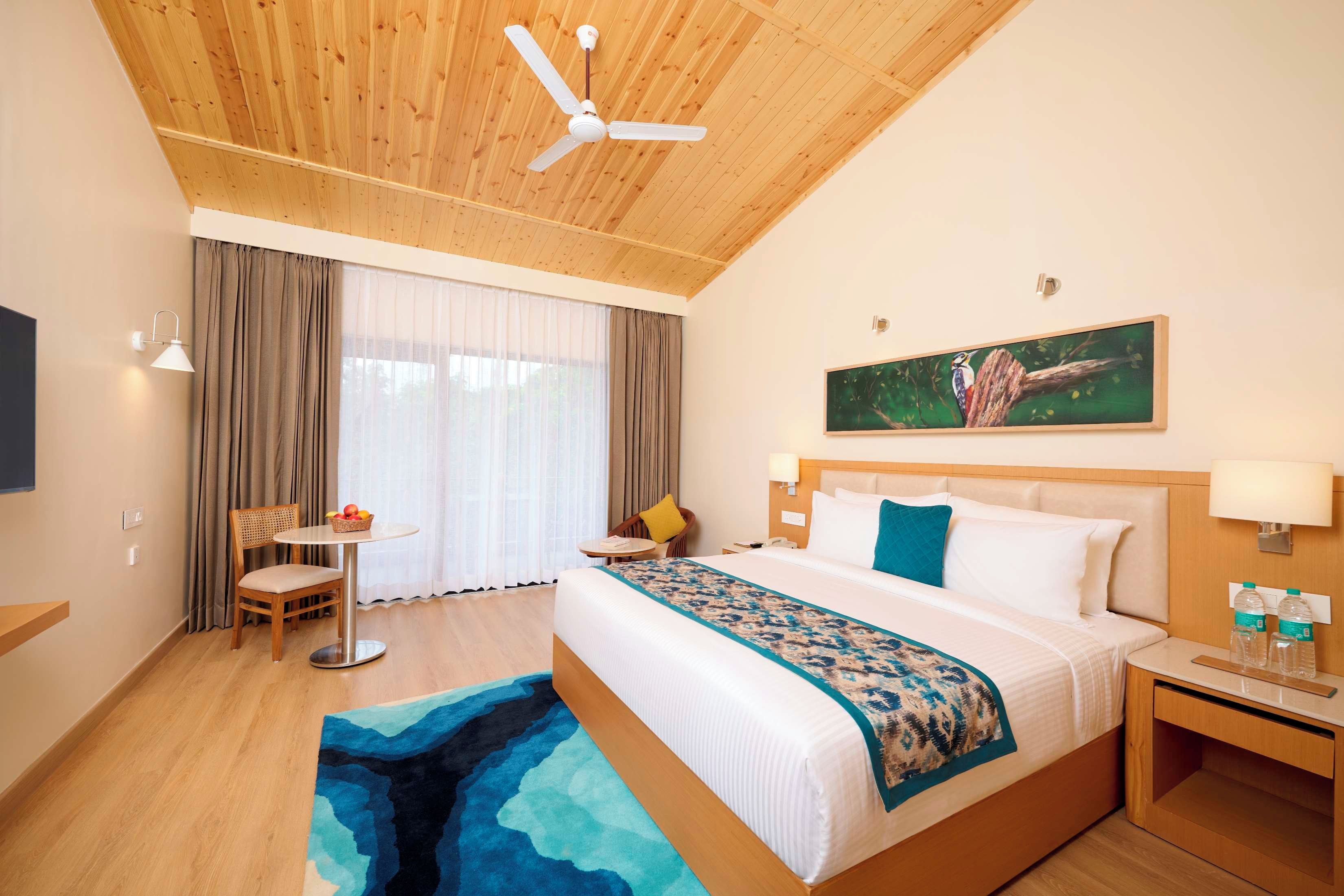 Quarto de hotel com cama, escrivaninha, cadeira, janela com cortinas, ventoinha no teto e um quadro na parede.