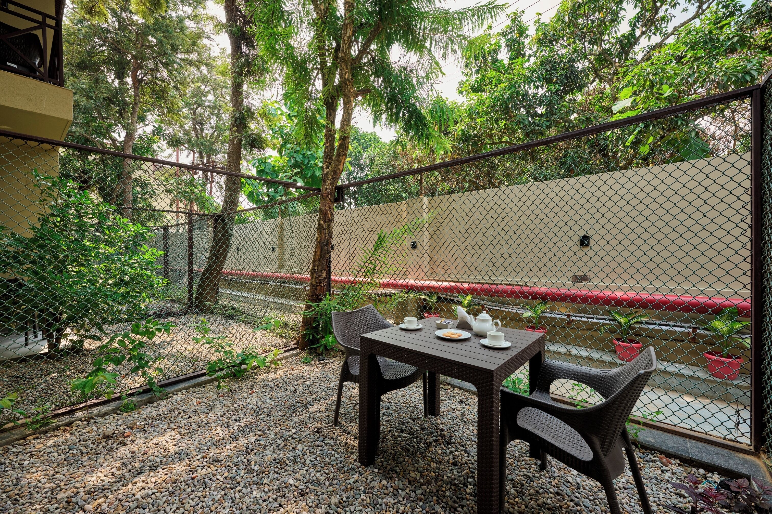 A patio area with a table and chairs, surrounded by a chain-link fence, with a view of a building and trees in the background.