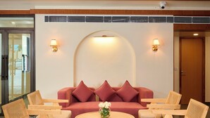 A lobby with a round wooden coffee table, a red sofa with cushions, and wooden chairs with woven backs. - ABN Sarovar Portico Jim Corbett  (Ramnagar)