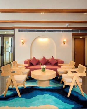 A lobby with a round wooden coffee table, a red sofa with cushions, and wooden chairs with woven backs. - ABN Sarovar Portico Jim Corbett  (Ramnagar)