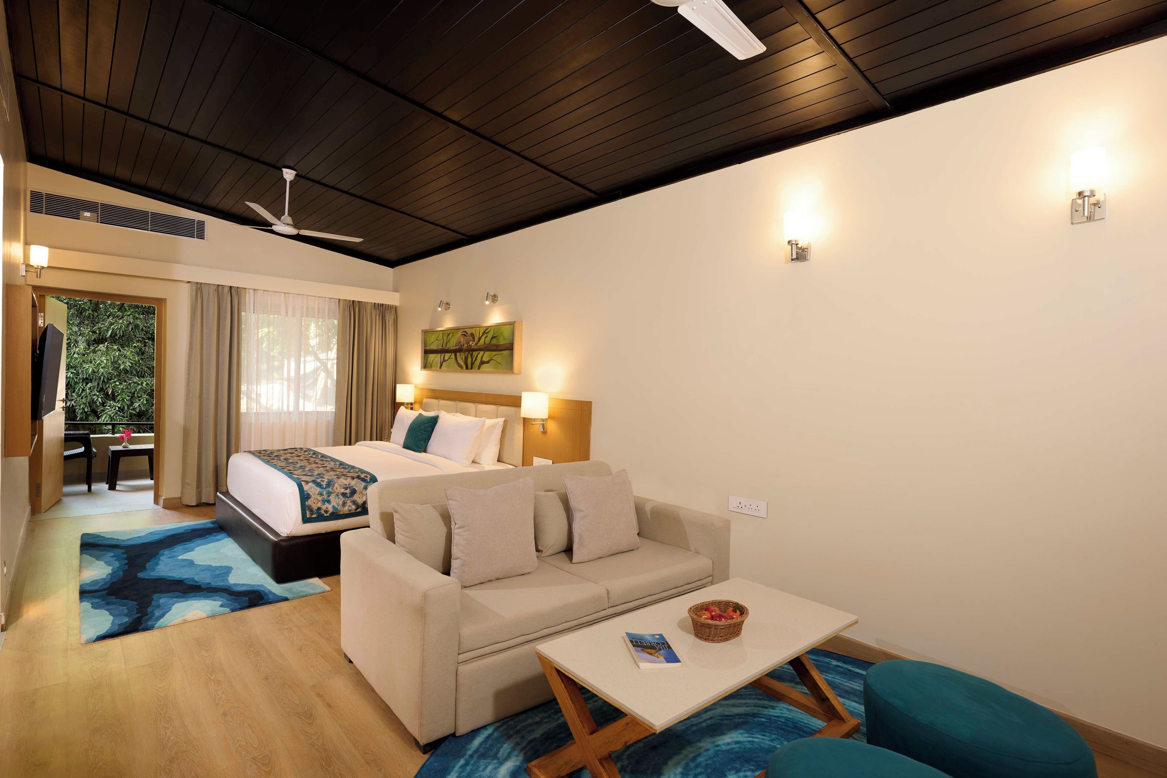 Une chambre d’hôtel avec un lit, un canapé, une table basse et une table à manger. On y trouve un ventilateur de plafond, une fenêtre avec des rideaux et un tableau au mur.