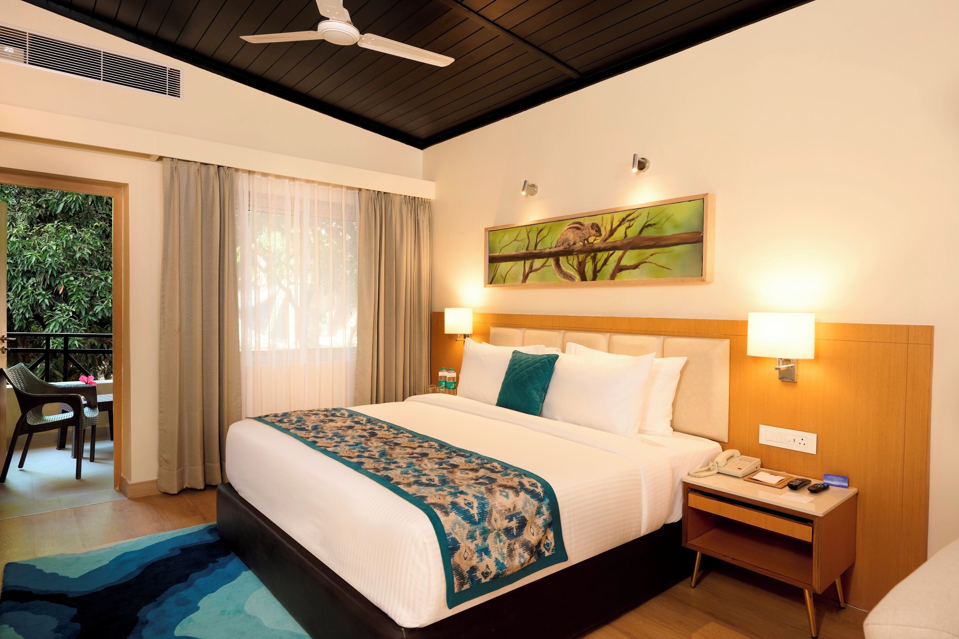 A hotel room with a bed, bedside lamps, a nightstand, a painting, and a view of greenery outside.
