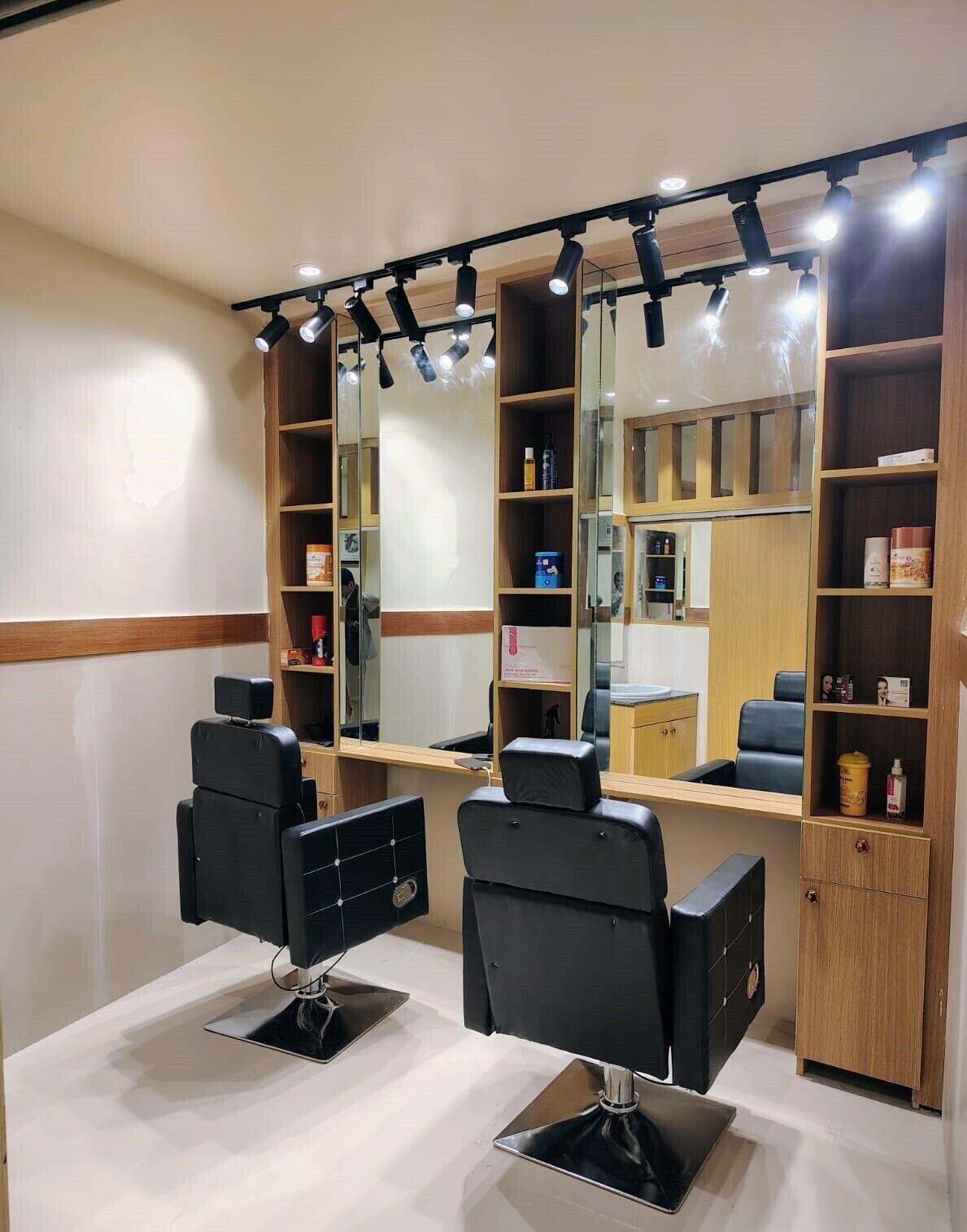 Un salón de belleza con dos sillas de peluquería negras, un espejo, estantes con productos para el cabello y un armario de madera.