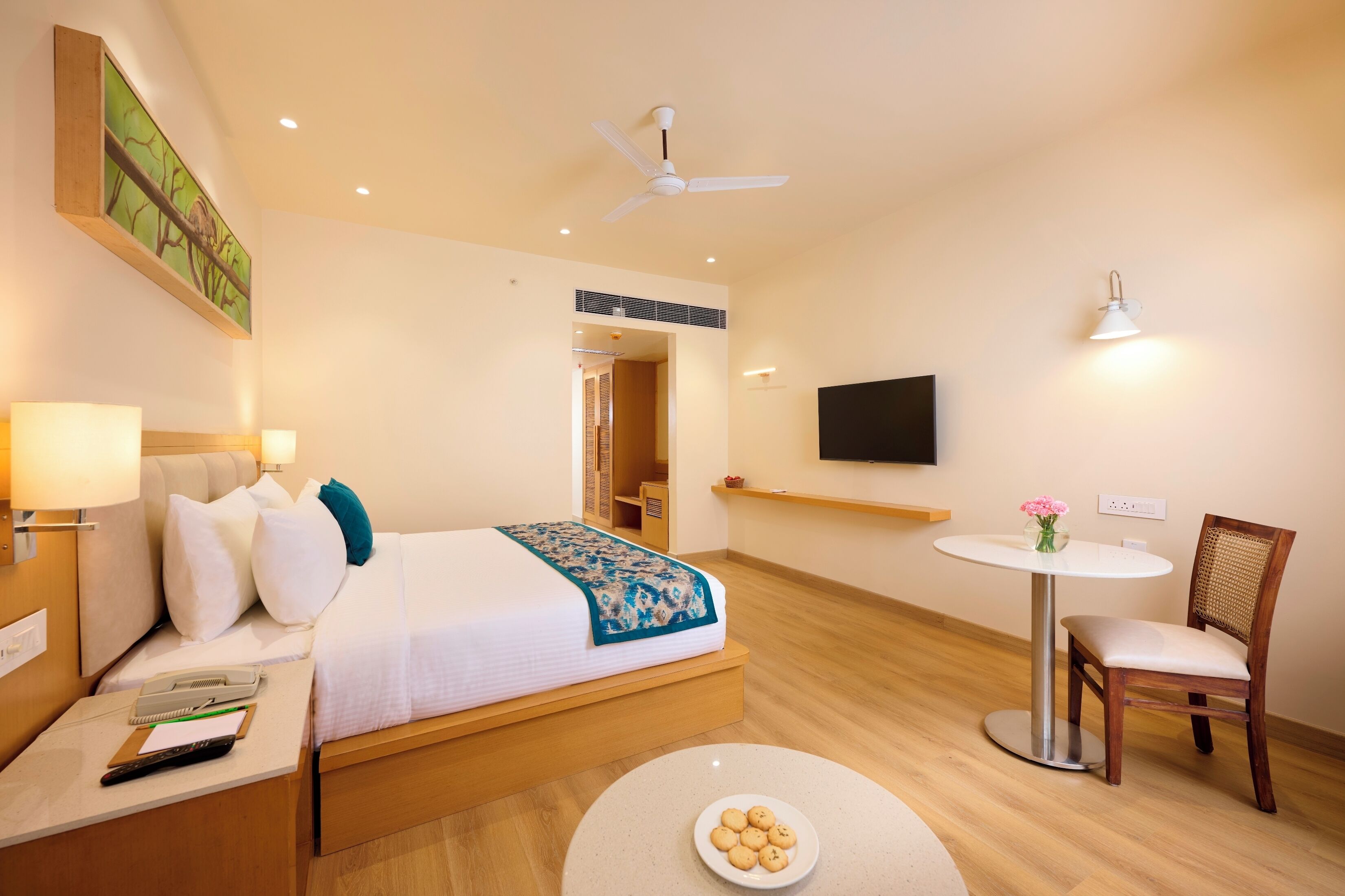 Una habitación de hotel con cama, mesita de noche, lámpara, silla, mesa redonda y ventilador de techo.