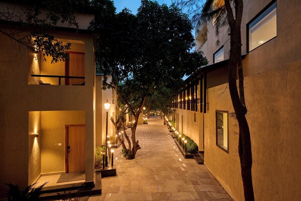 A narrow, well-lit corridor with a wooden door at the end, flanked by buildings with windows and outdoor lighting. - ABN Sarovar Portico Jim Corbett  (Ramnagar)