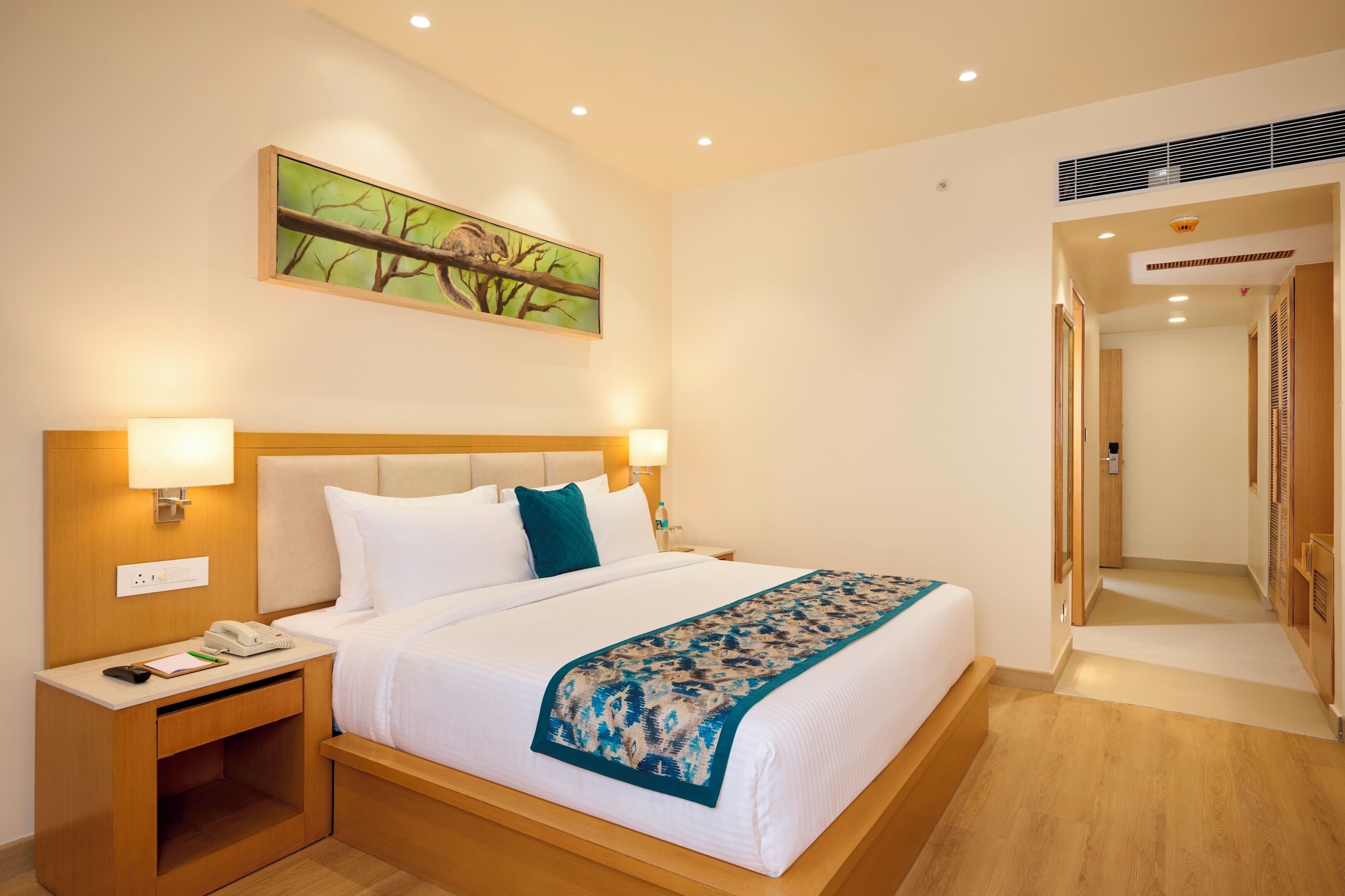 Habitación de hotel con una cama grande, dos lámparas de noche, una mesita de noche con teléfono y bloc de notas, un cuadro en la pared y un pasillo con puertas de madera.