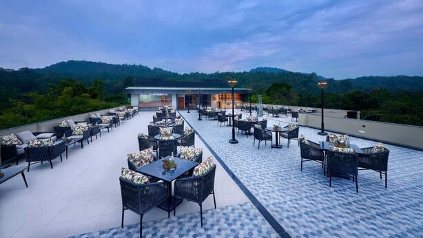 An outdoor patio with tables and chairs, a building in the background, and a view of a forested hill. - ABN Sarovar Portico Jim Corbett  (Ramnagar)