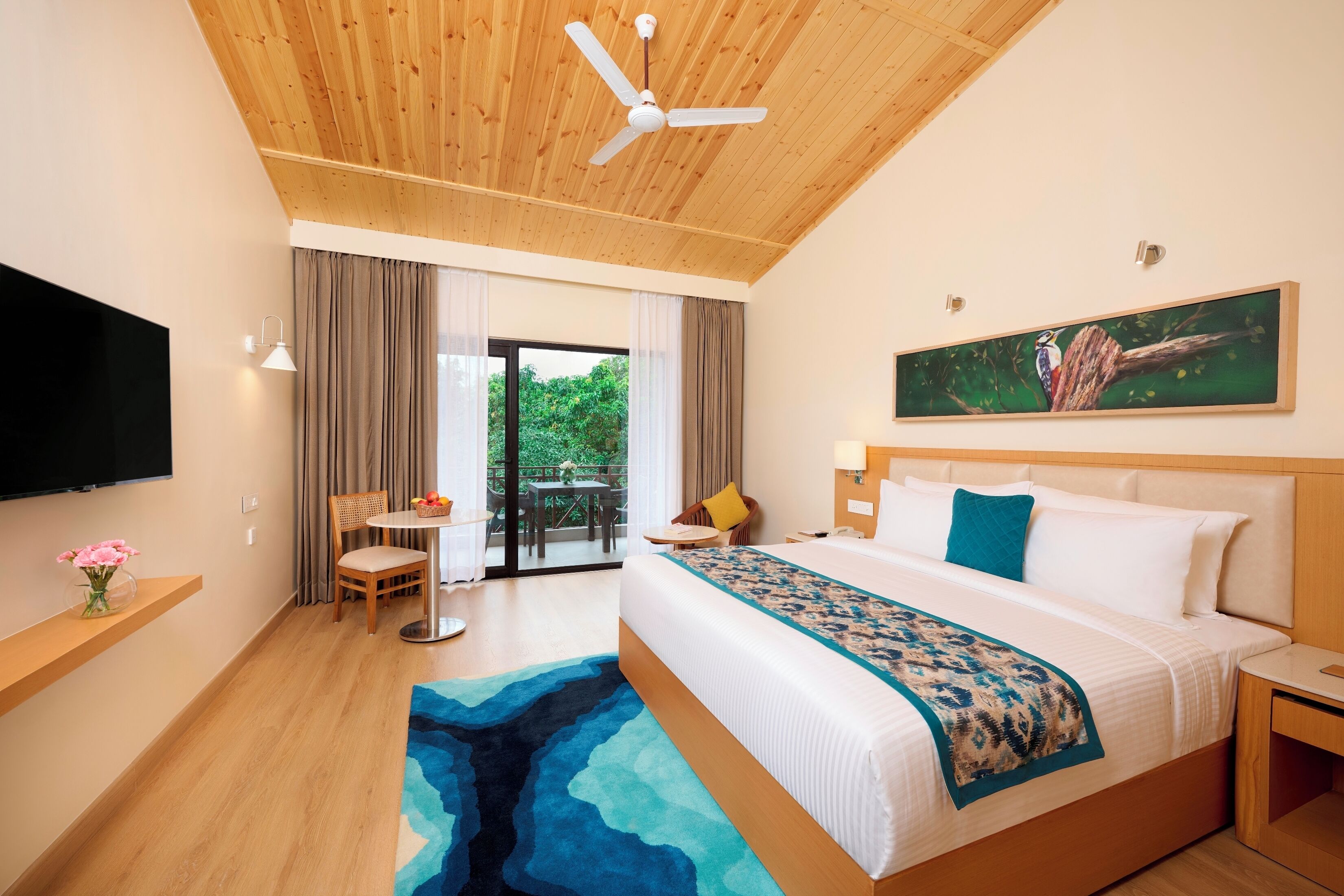 Camera d'albergo con un letto, una ventola a soffitto, un quadro, un televisore, una finestra con tende e un tavolino con sedie.