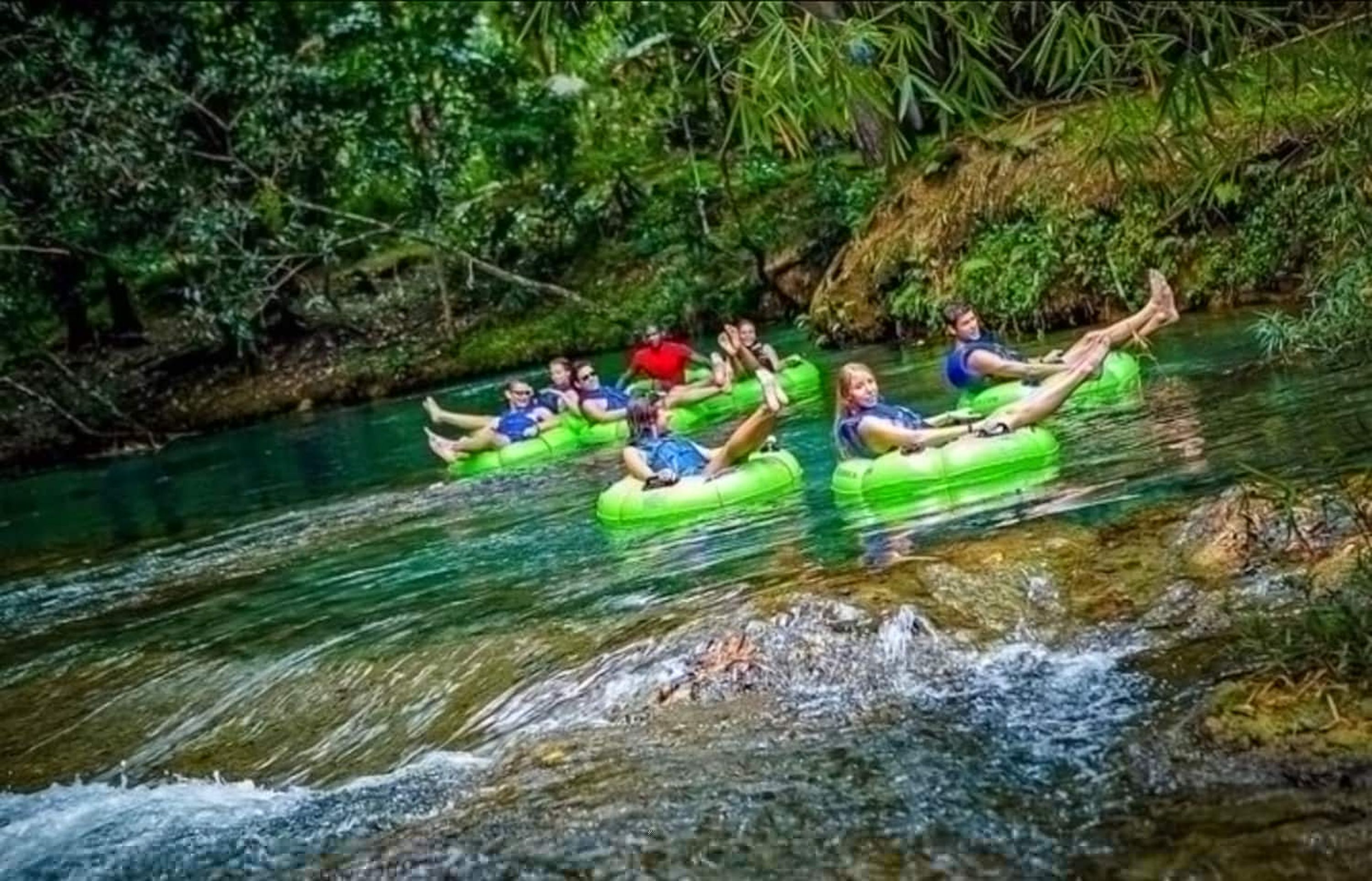 People floating on green inflatable tubes in a river surrounded by dense greenery.