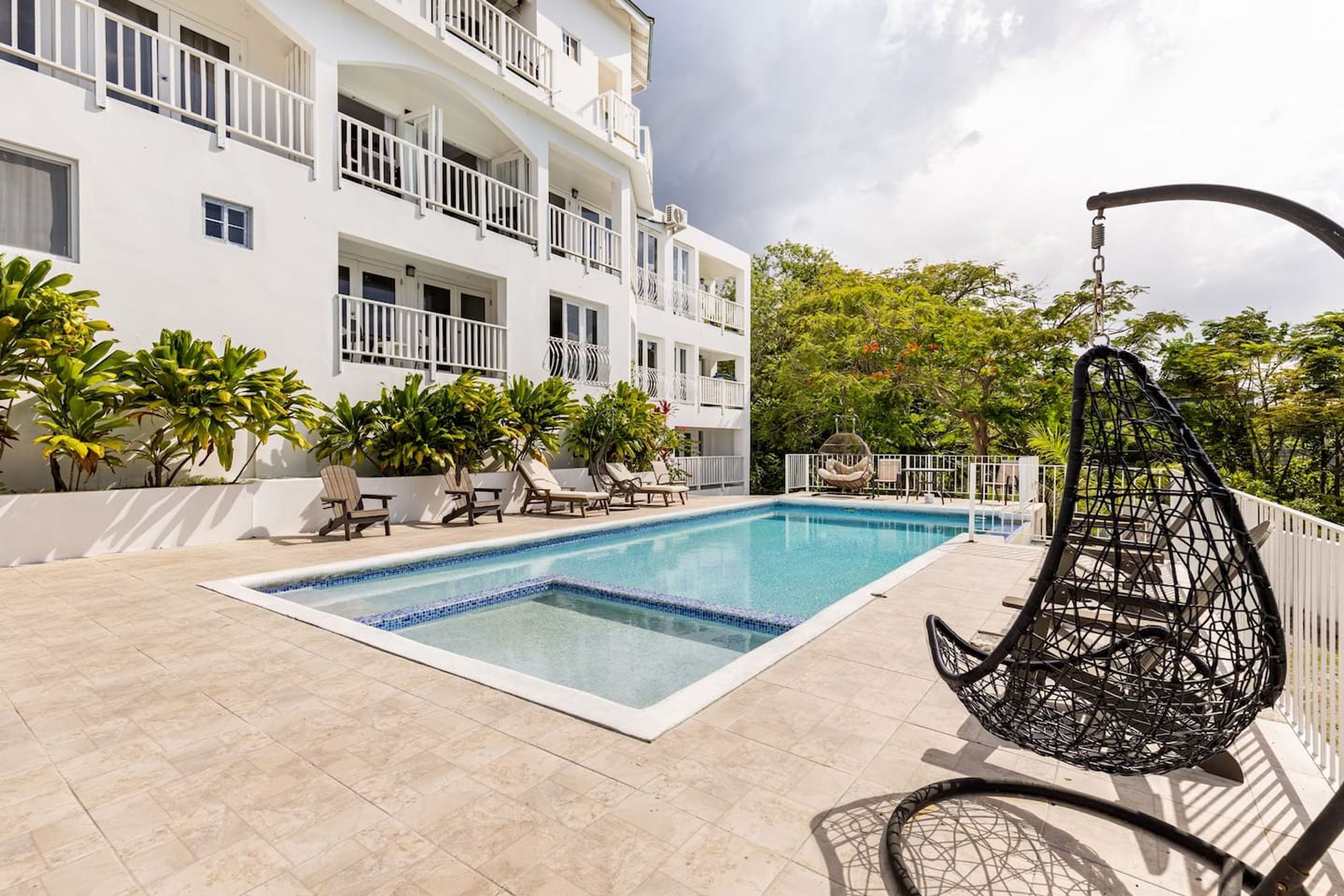 Une piscine entourée de chaises longues et d’un fauteuil suspendu, le tout adossé à un bâtiment blanc doté de balcons et d’une végétation luxuriante.