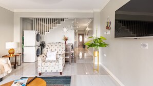 Sala de estar moderna com sofá, mesa de centro e área de jantar com cadeiras. Uma escada leva a uma área de cozinha com geladeira e máquina de lavar.