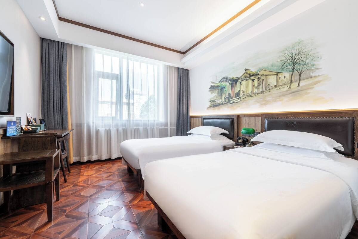 A hotel room with two beds, a desk, and a large window with curtains.