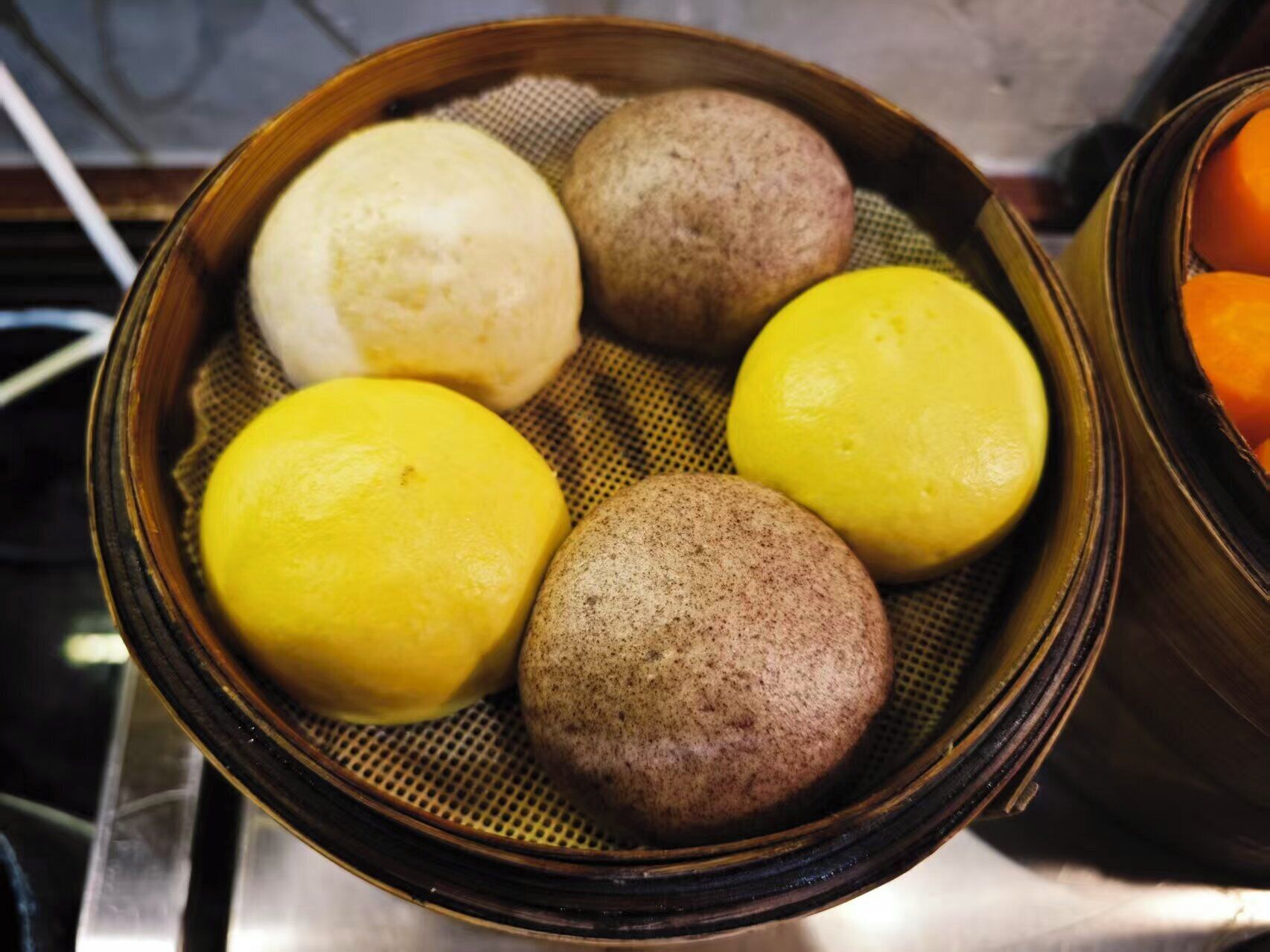 A bamboo steamer containing five round, steamed buns in different colors.