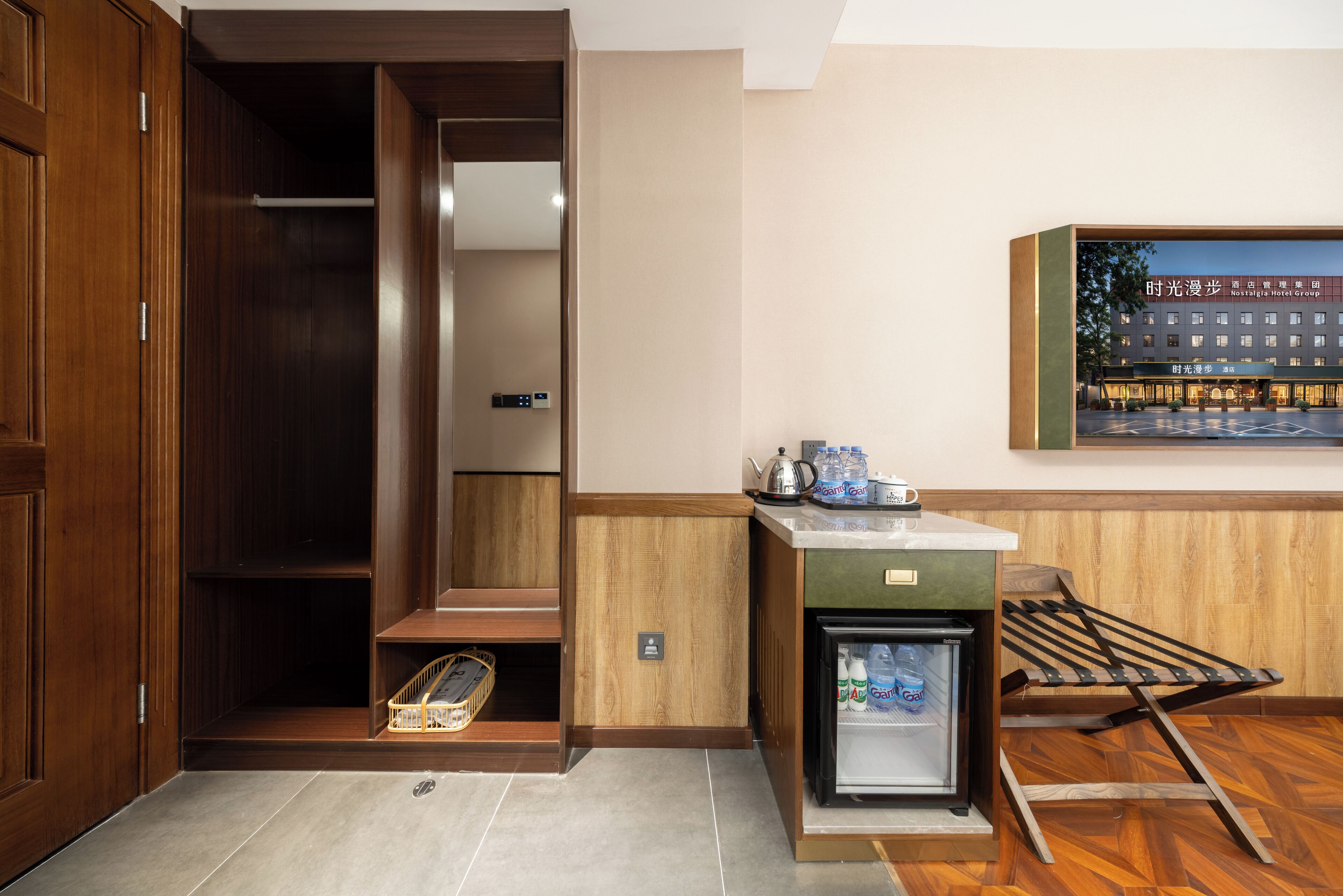 A hotel room with a wooden wardrobe, a small kitchenette with a refrigerator, a sink, and a microwave. There is a wooden bench and a small table with a kettle and cups.