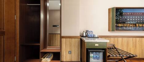 A hotel room with a wooden wardrobe, a small kitchenette with a refrigerator, a sink, and a microwave. There is a wooden bench and a small table with a kettle and cups.