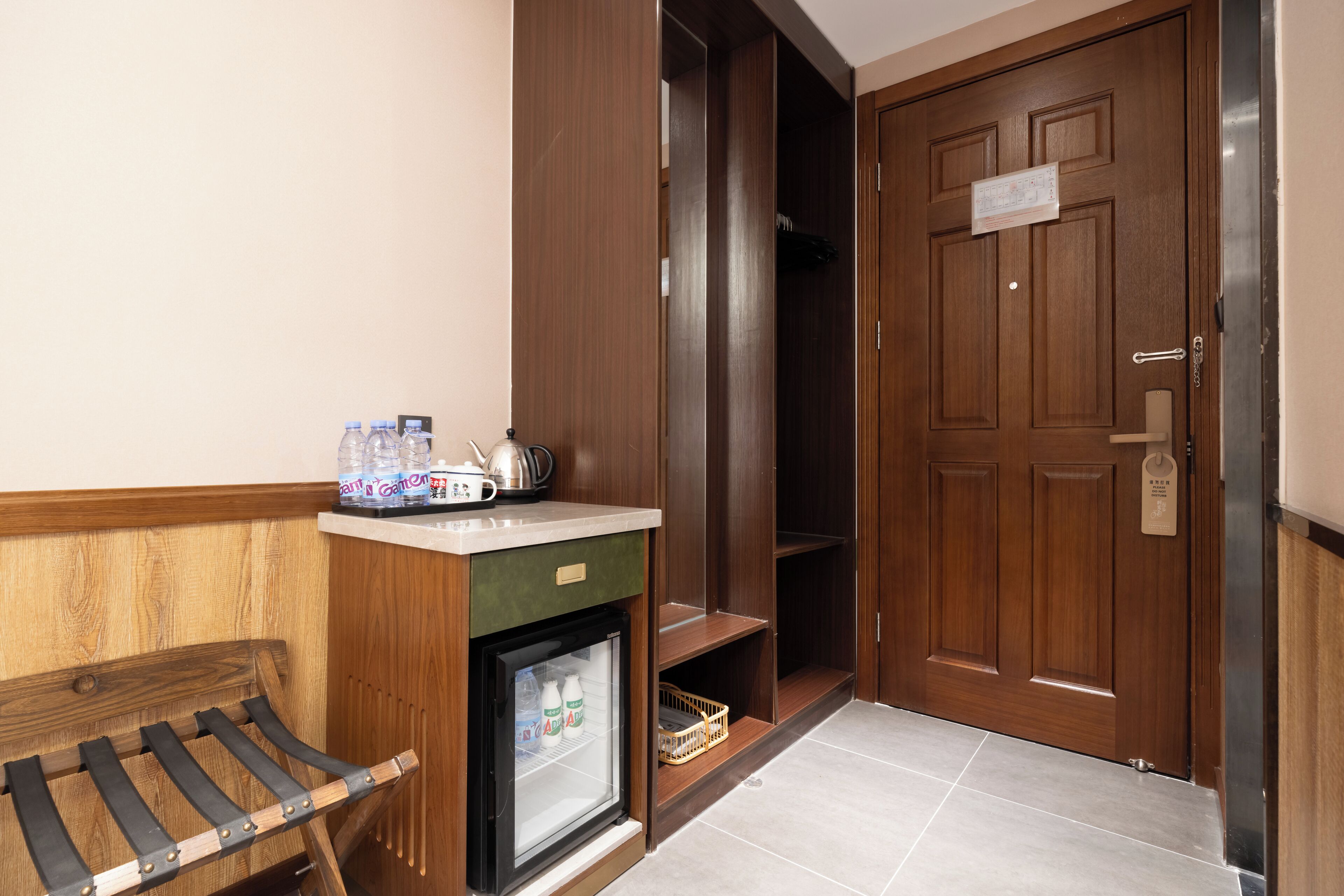 Ein Hotelzimmer mit einer hölzernen Bank, einer Minibar, einer Tür mit einem Schild und einem Fliesenboden.