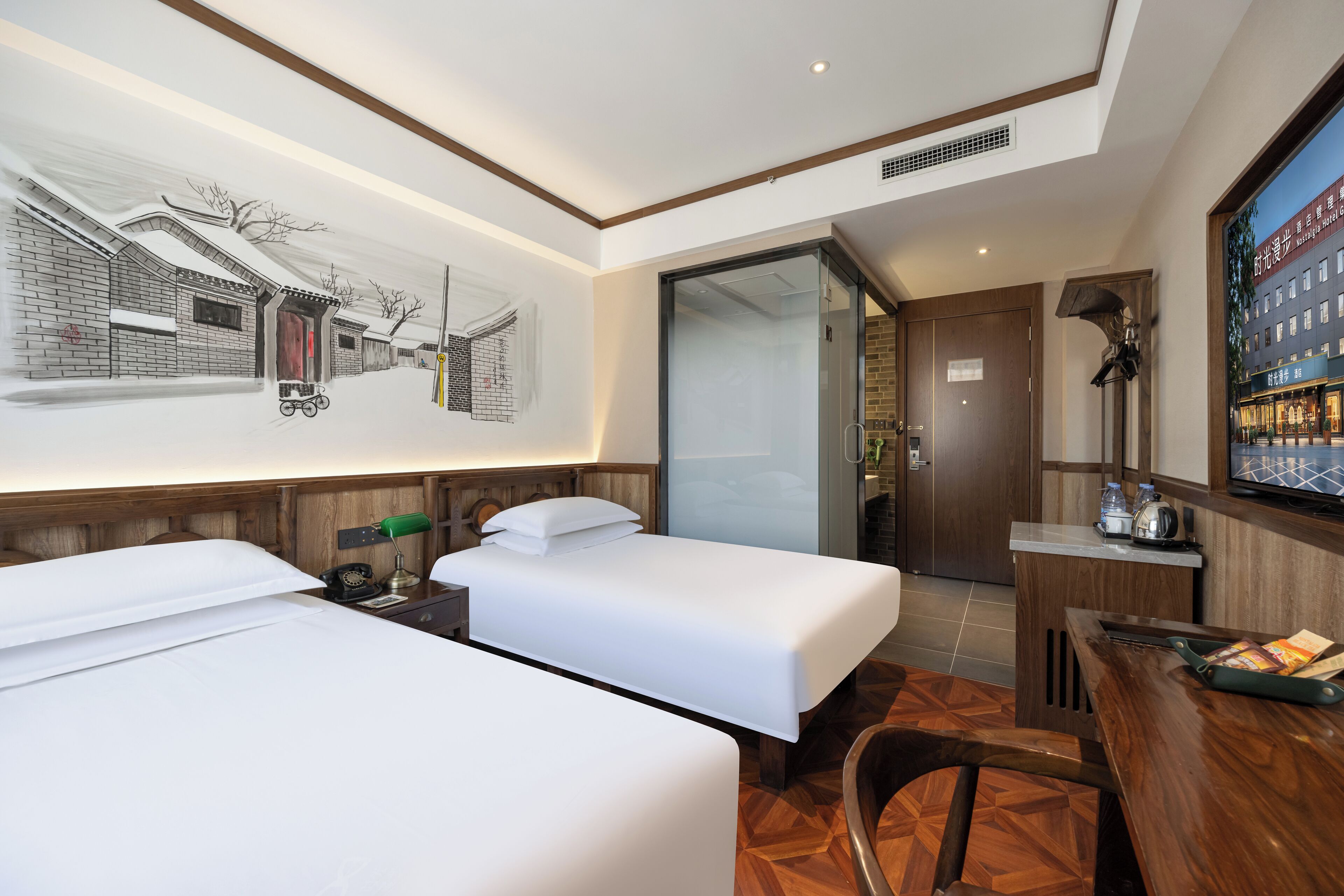 A hotel room with two single beds, a desk with a chair, a small table, a lamp, and a wall-mounted TV.