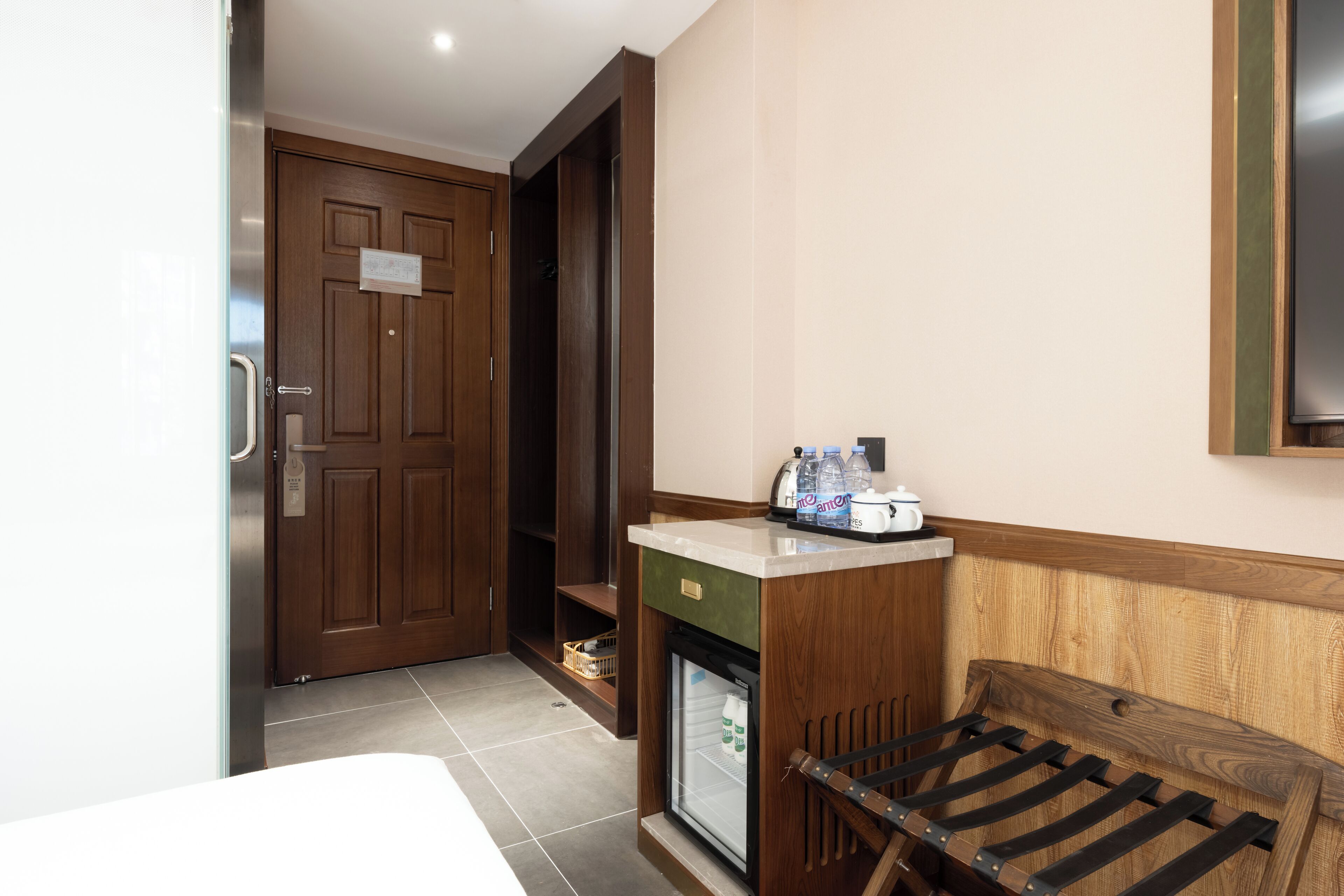 Habitación de hotel con puerta de madera, un minibar, un refrigerador pequeño, una banca de madera y un televisor instalado en la pared.
