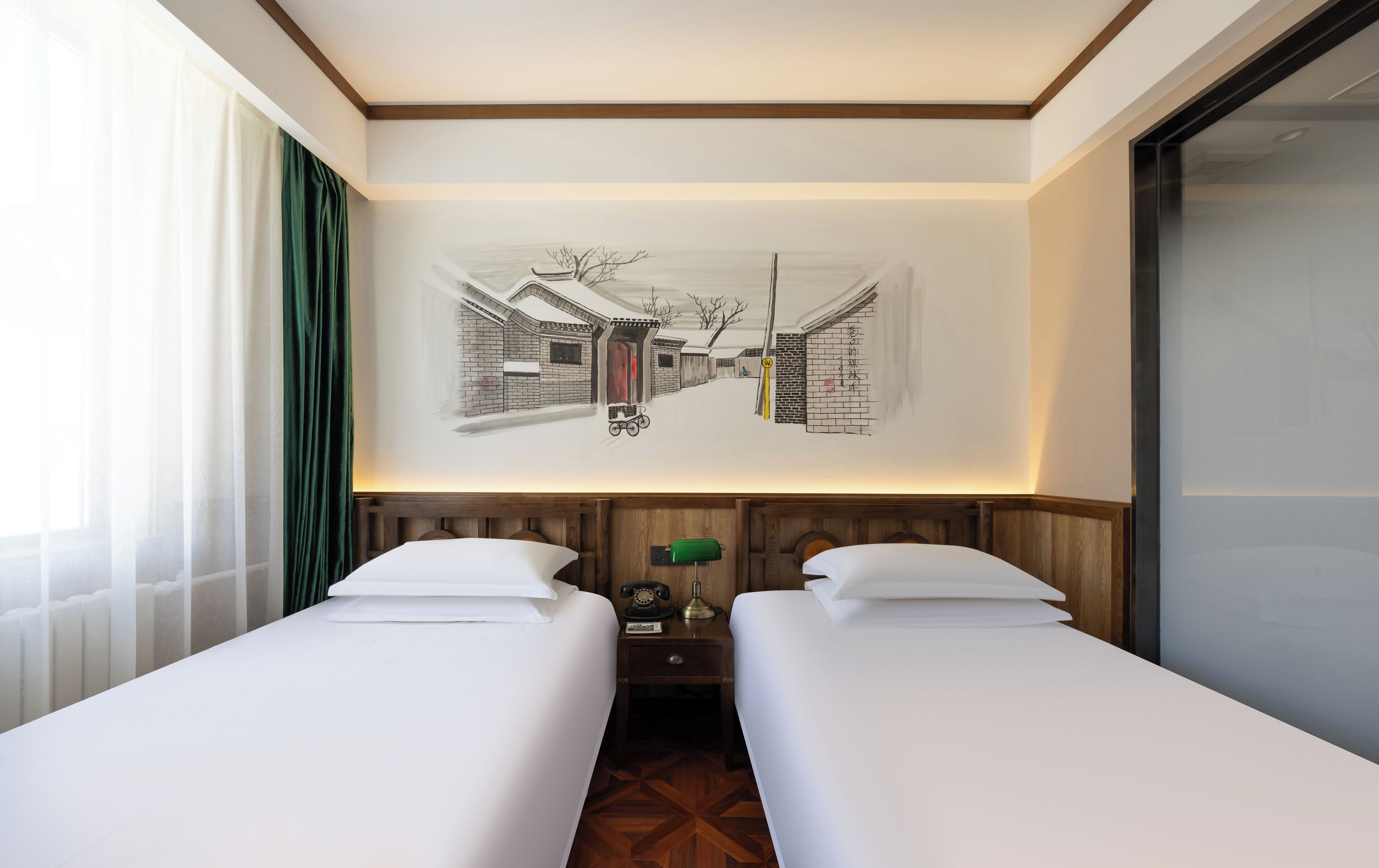 A hotel room with two single beds, a wooden headboard, a wall-mounted artwork depicting a snowy street scene, and a window with sheer curtains.