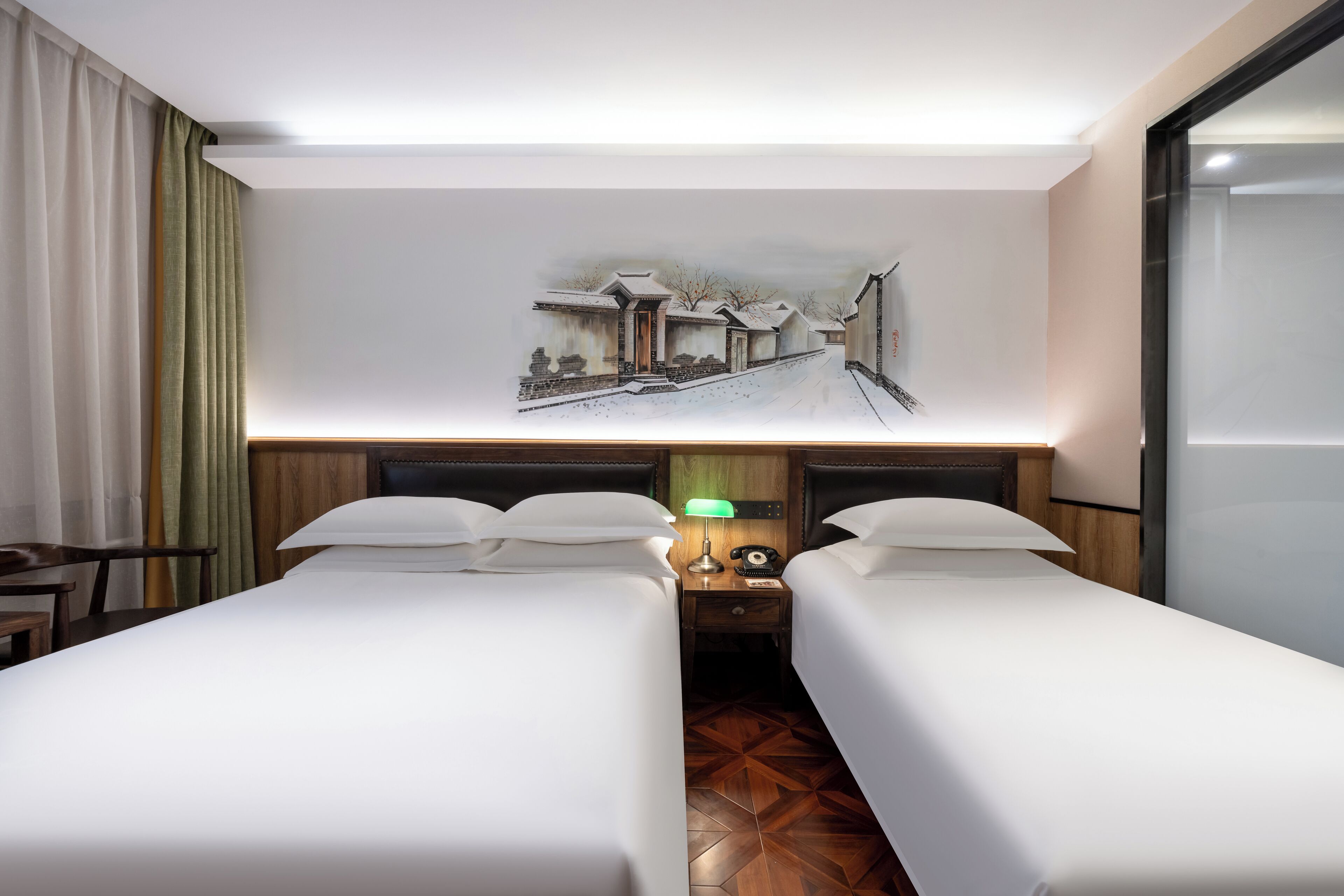 A hotel room with two beds, a nightstand with a lamp, a painting on the wall, and a wooden floor.
