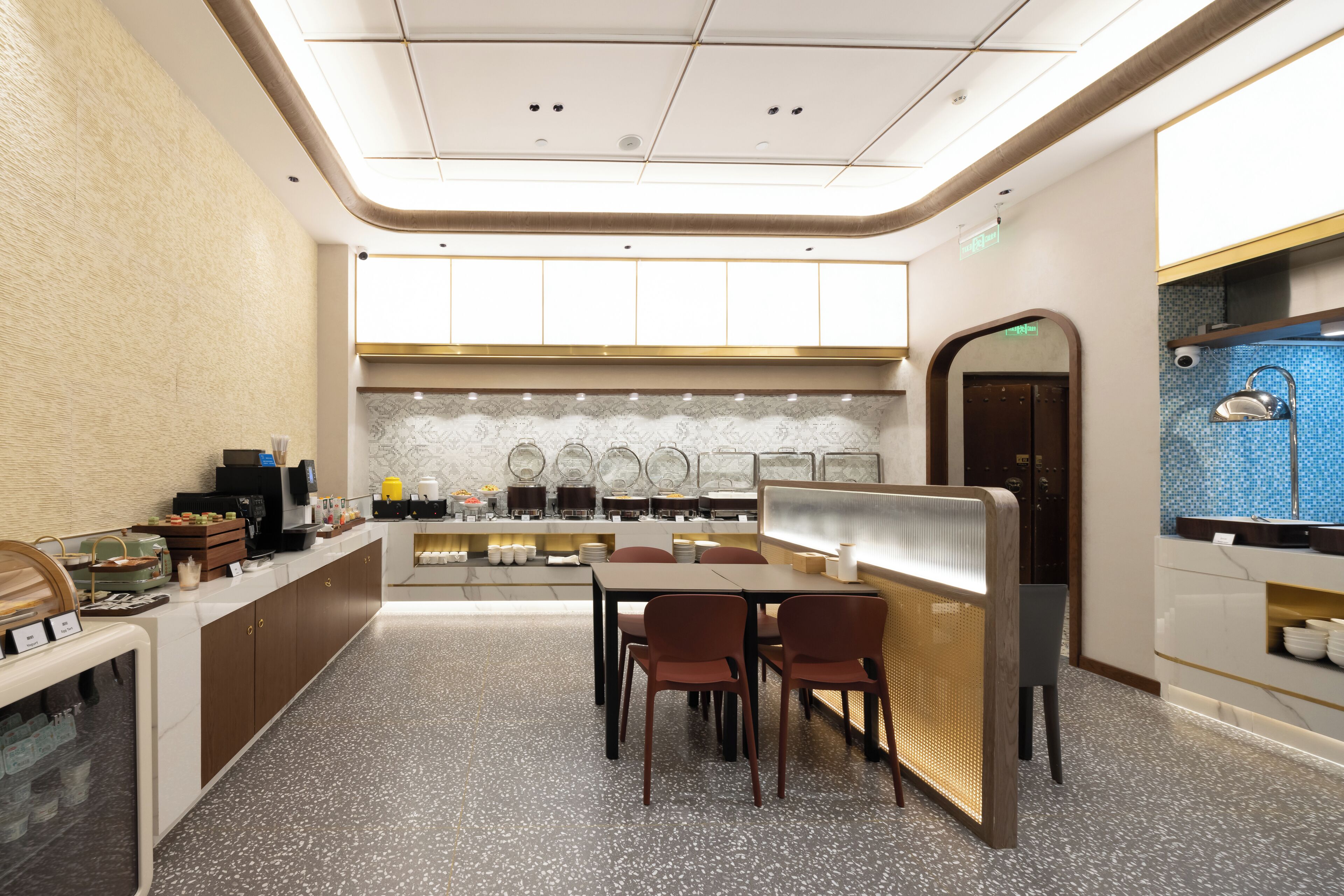 A modern hotel lobby with a central seating area, a reception desk, and a kitchen area in the background.