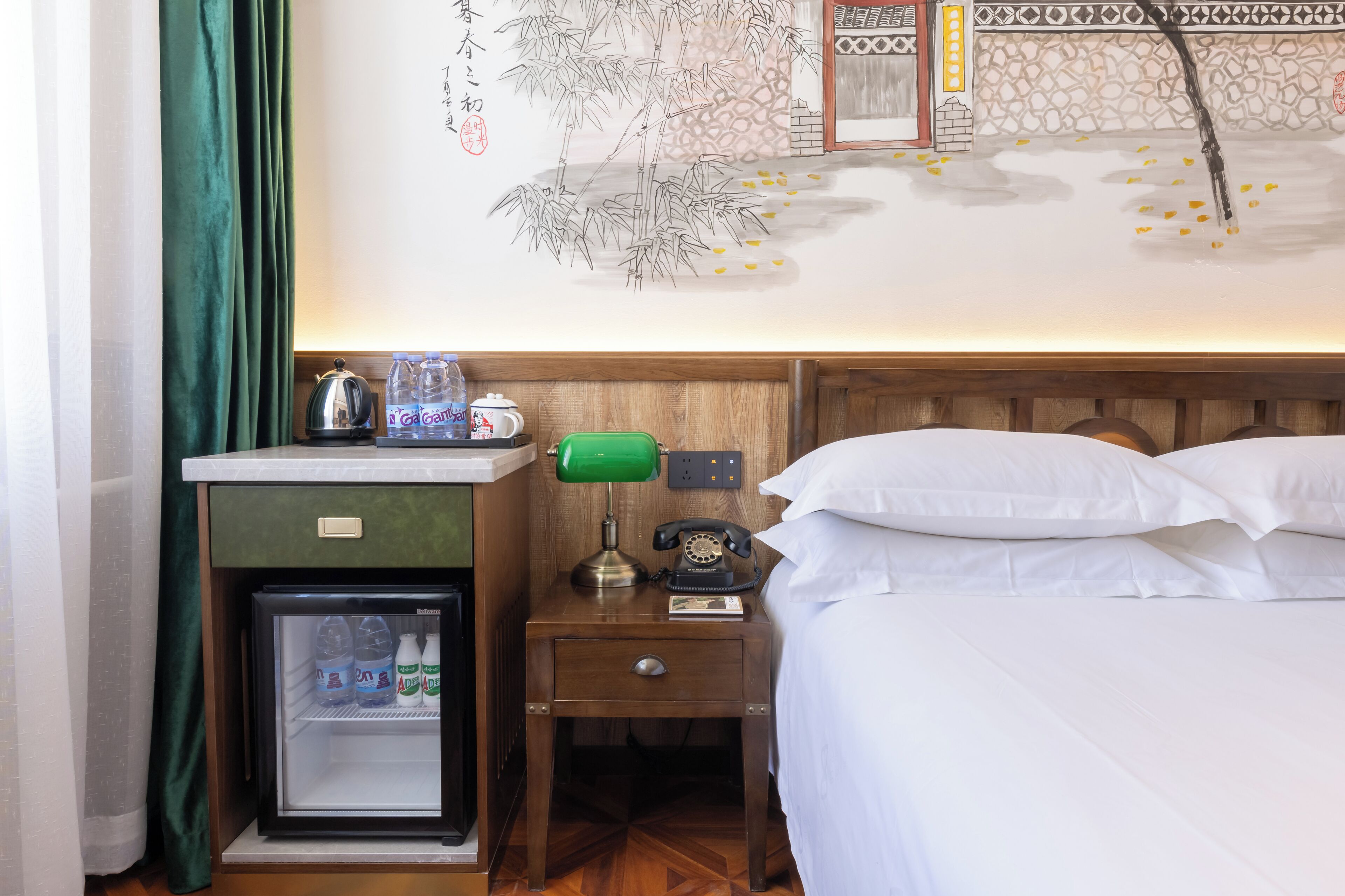 A hotel room with a bed, a nightstand with a lamp, a refrigerator, and a wall with a decorative mural.