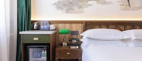 A hotel room with a bed, a nightstand with a lamp, a refrigerator, and a wall with a decorative mural.