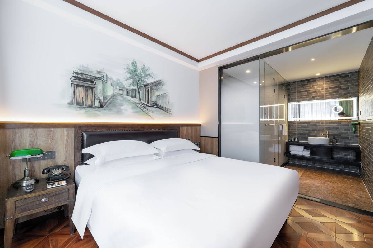 A modern hotel room with a large bed, a nightstand with a lamp, a telephone, and a painting on the wall.