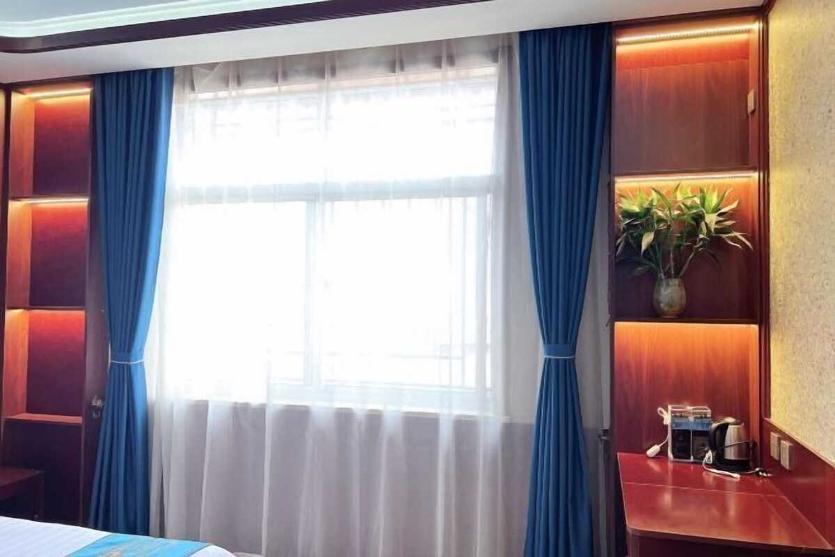 Quarto de hotel com uma janela grande, cortinas, uma mesa de cabeceira com cafeteira e uma planta em uma estante.