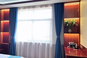 Quarto de hotel com uma janela grande, cortinas, uma mesa de cabeceira com cafeteira e uma planta em uma estante.
