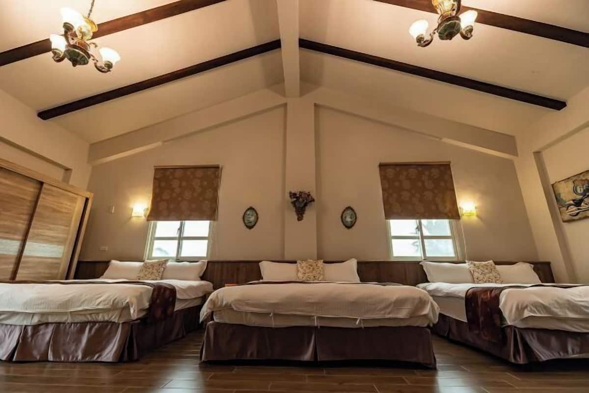 A large room with three beds aligned in the center, wooden beams on the ceiling, and wall-mounted light fixtures.