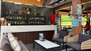 Un espace bar avec des étagères remplies de bouteilles d’alcool, un réfrigérateur pour les boissons Coca-Cola et un coin salon avec des meubles en osier.